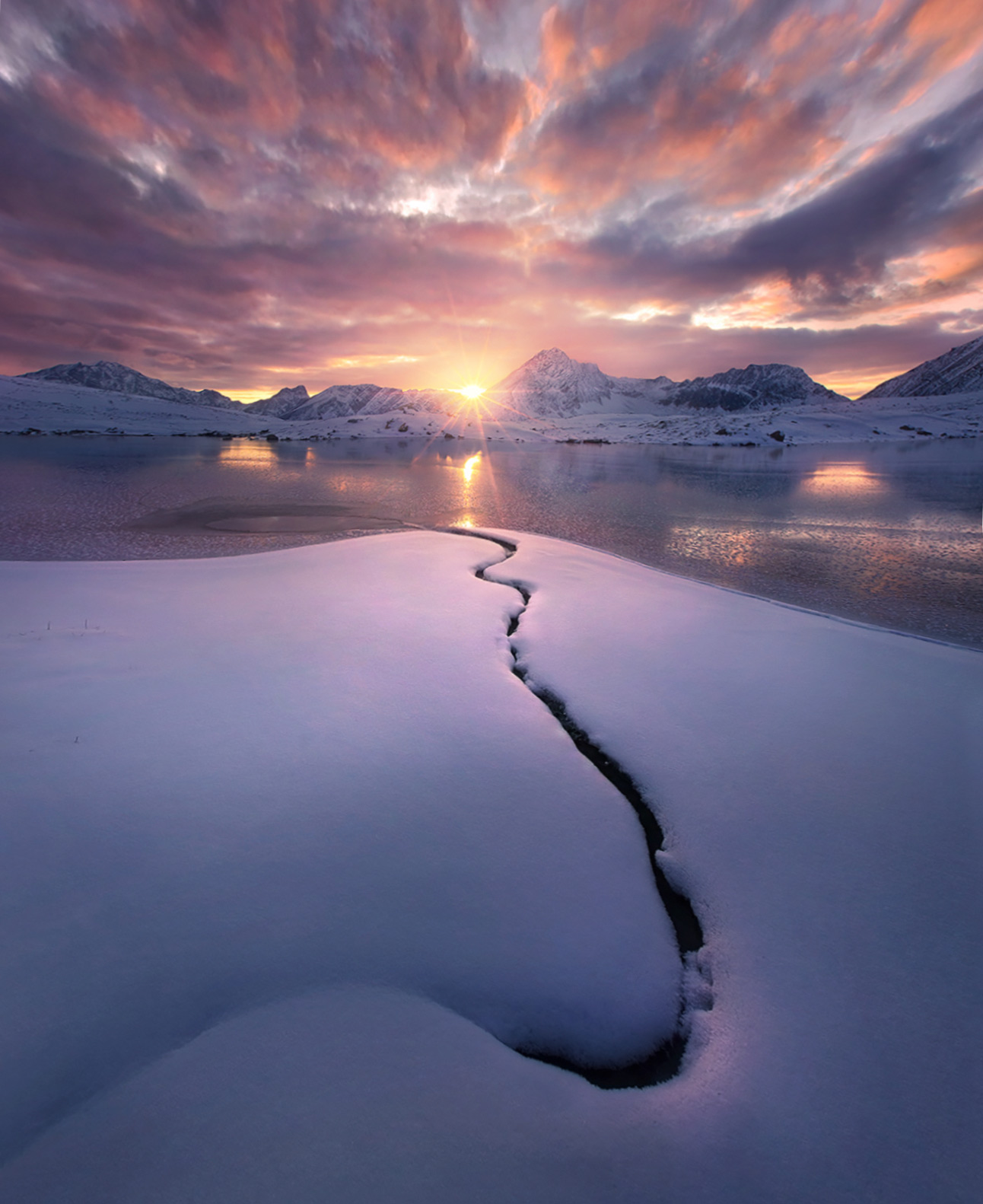 The solitary unfrozen line of a burried stream disappears into a frozen lake and the sunset beyond, photographed from a very...
