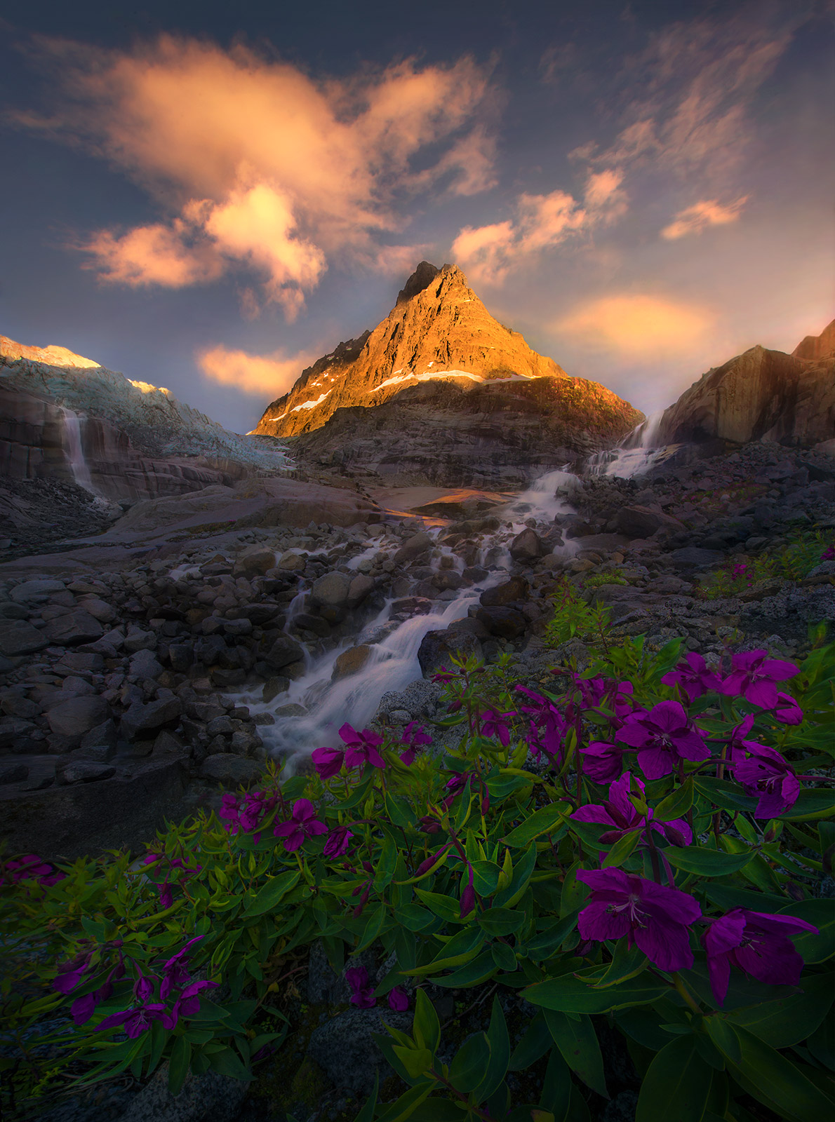 Lush summer wildflowers adorn the glacial hillside. &nbsp;Above, mountains reach into the sunset sky surrounded by high waterfalls...