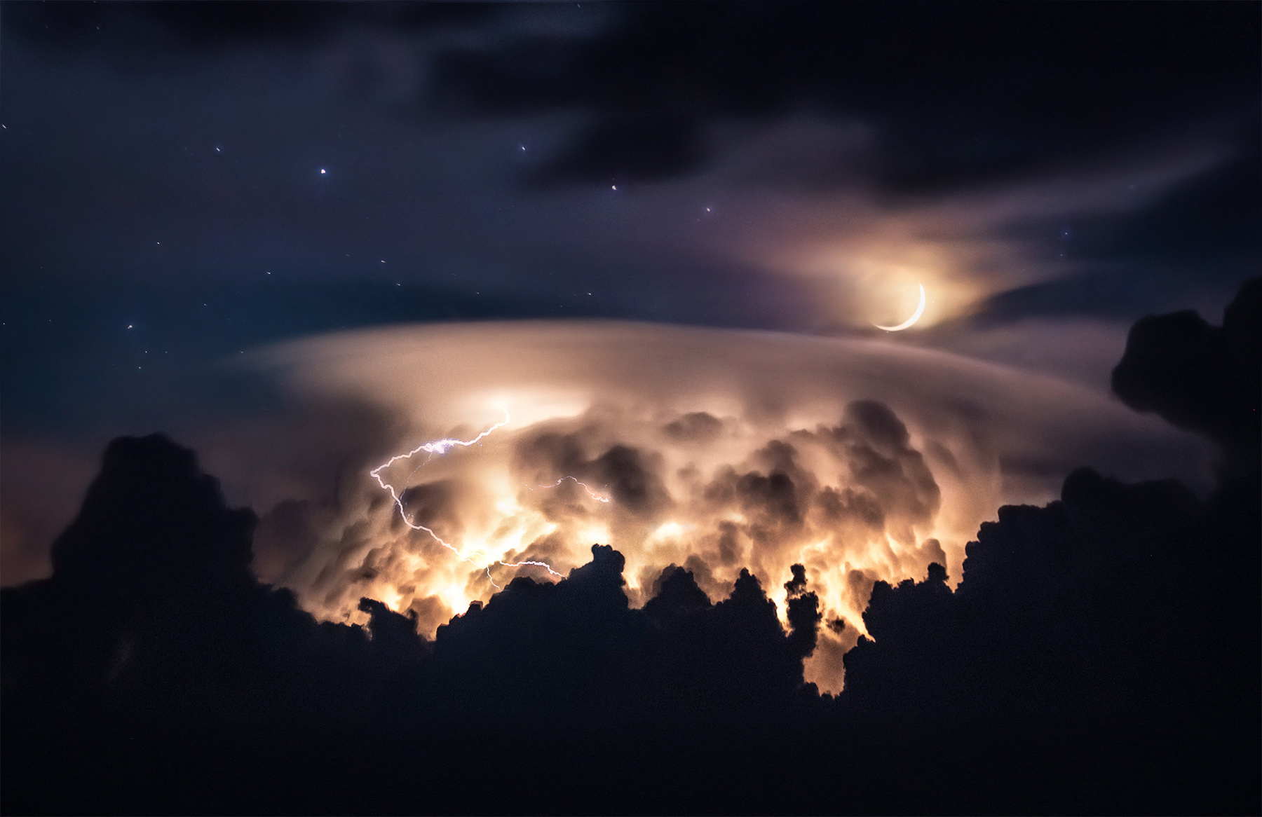 Layers of cloud tops illuminated by lightning next to the setting crescent moon at twilight through a long lens.