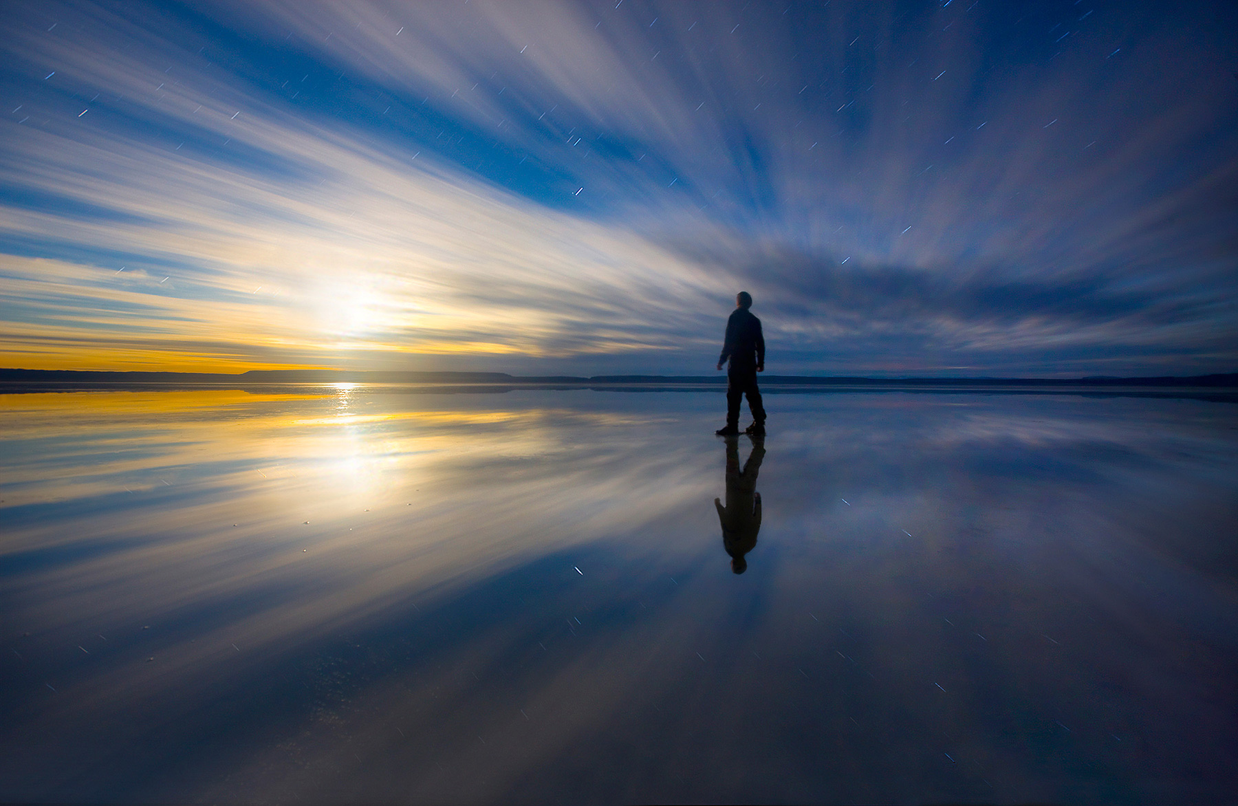 A lone figure stands still in shallow water on Oregon's Alvord playa amidst the surreal fusion of night sky, streaking clouds...