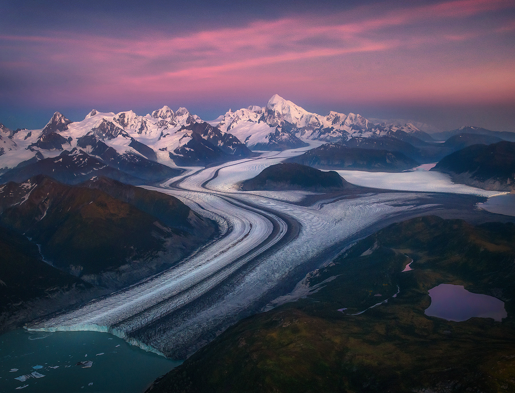 Enormous mountains and glaciers more than 20 miles long envelope an Alaskan landscape adorned with fall colors at twilight.